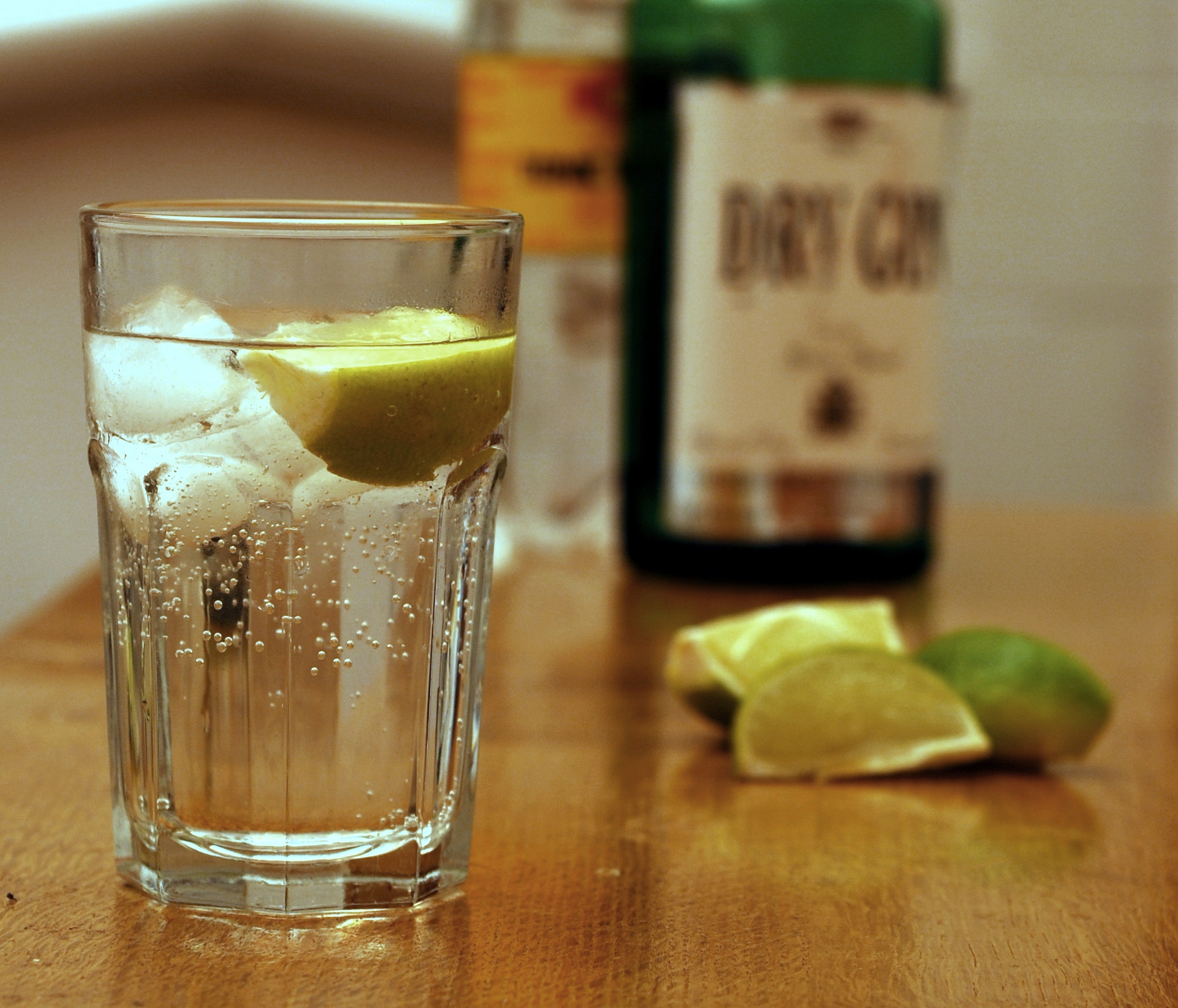 Photograph of a gin and tonic cocktail in a clear drinking glass with a wedge of lime floating in it. The liquid in the glass is clear with some bubbles. More wedges of lime and a bottle of gin and a bottle of tonic can be seen in the background.