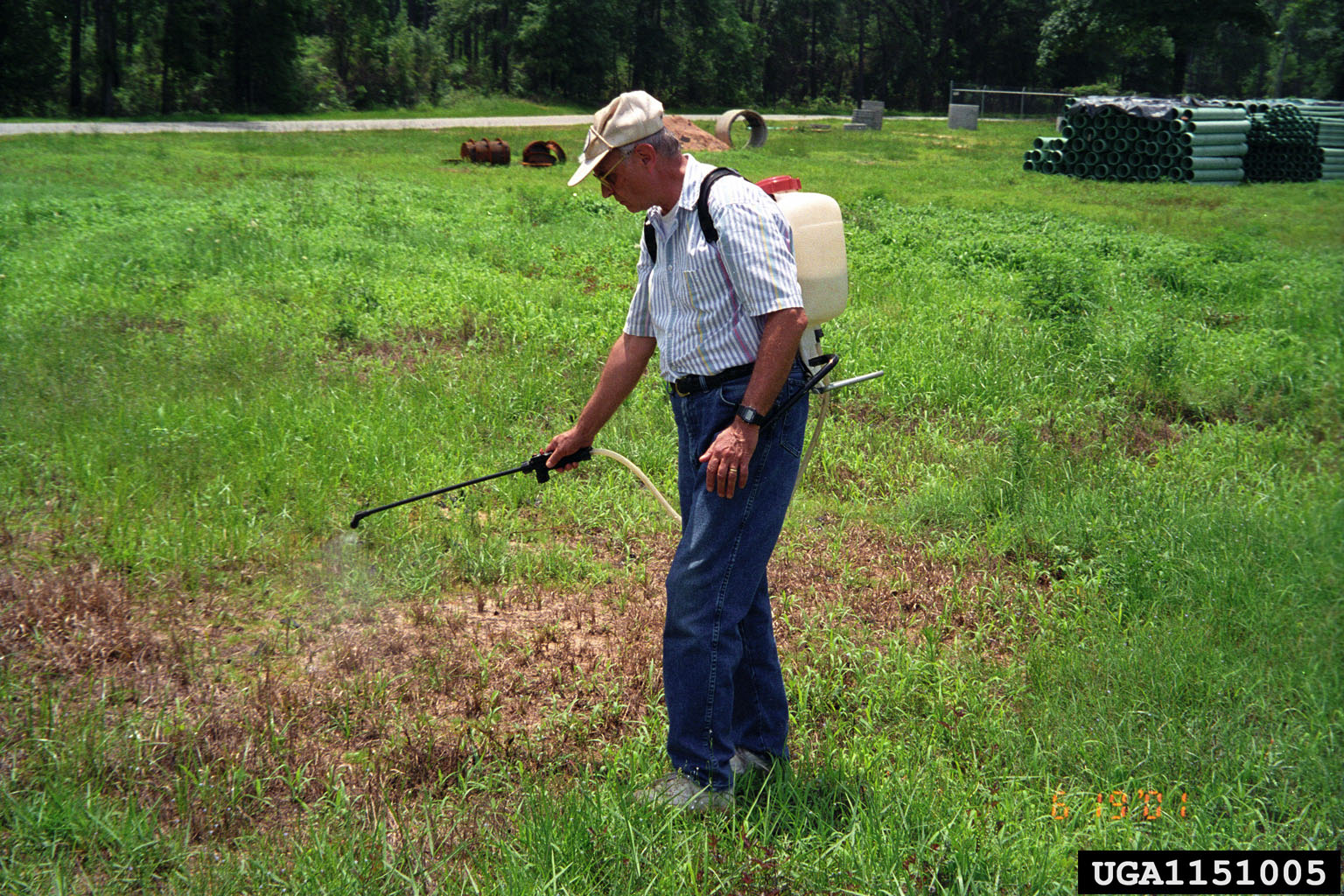 Photograph of a man in a white shirt and jeans and beige hat spraying herbicide onto a field of grass.