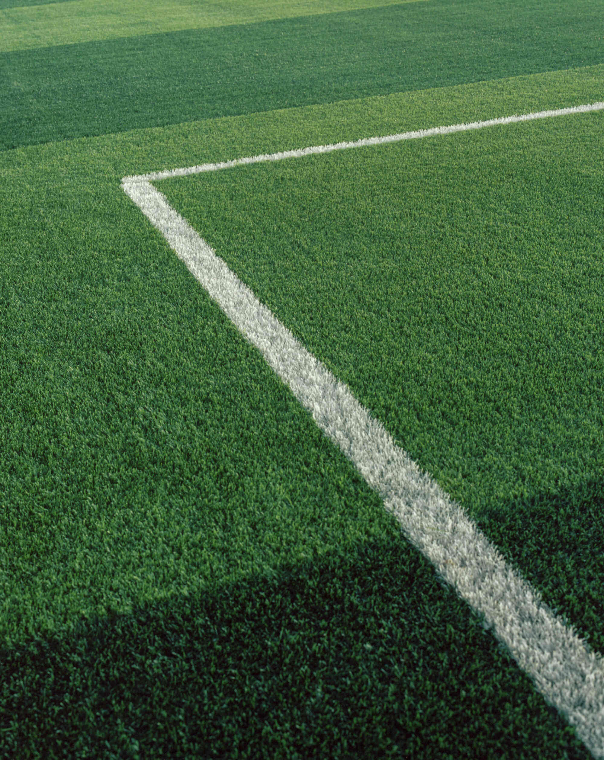 Photograph of synthetic turf on an athletic field. The photo shows green turf and one white line with a 90-degree angle in it.
