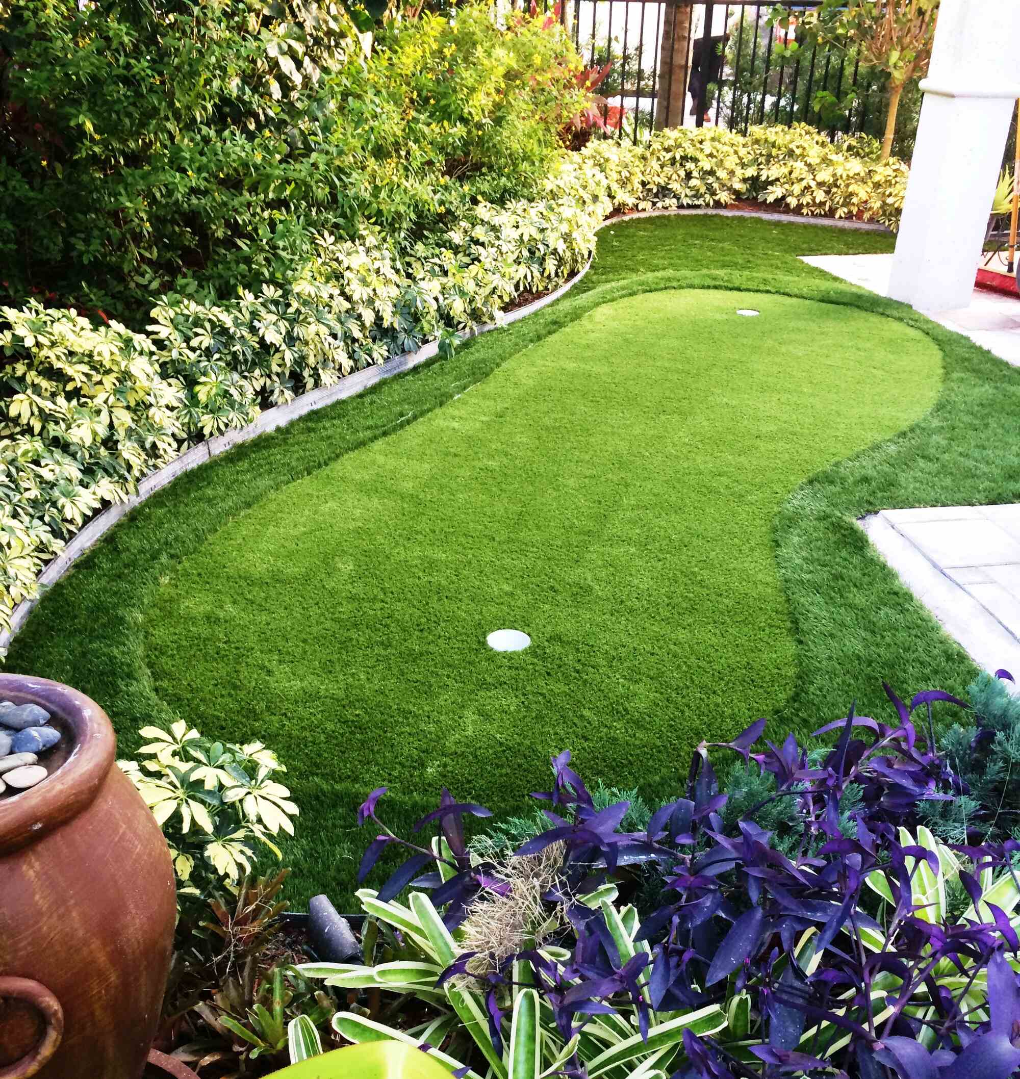 Photograph of a small putting green made of synthetic turf that appears to be near a residence. The putting green is roughly oval with two holes in it.