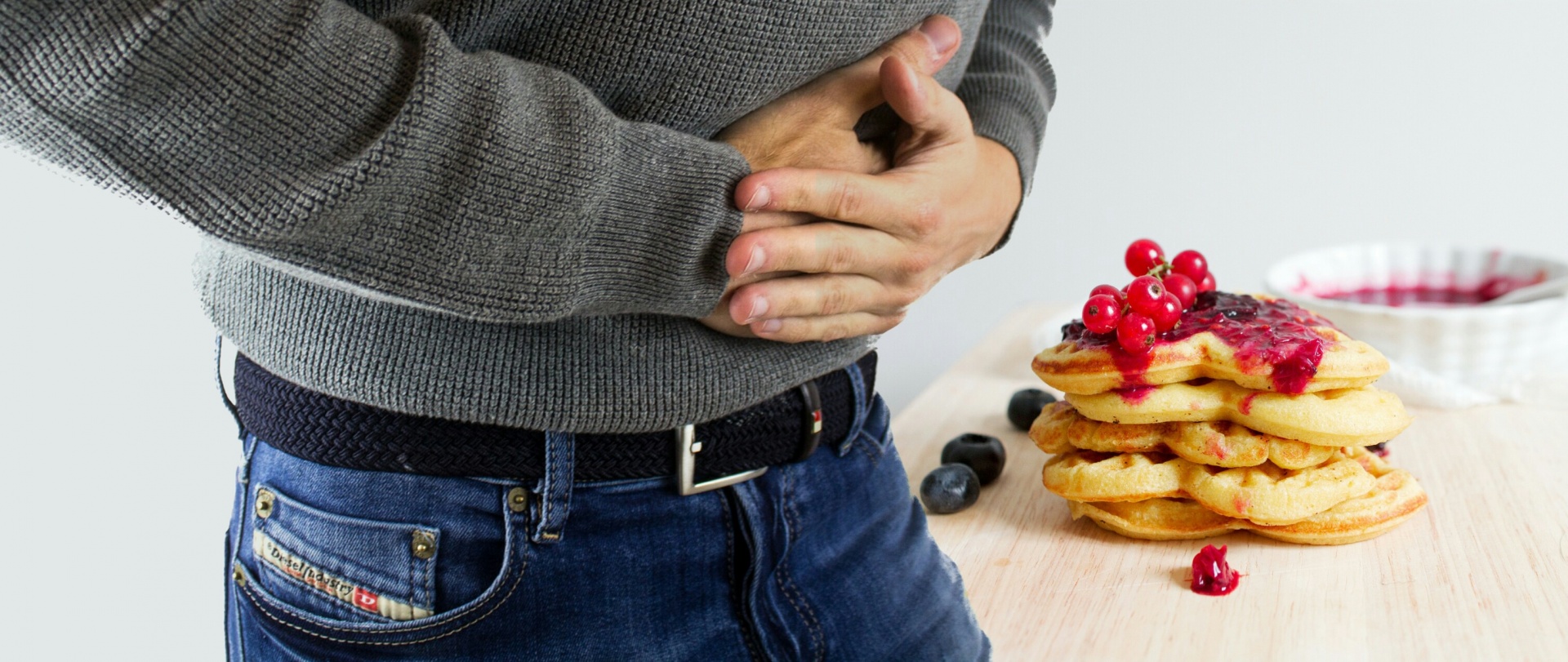 Photograph of a person clasping his hands over his stomach, indicating that he is in pain. A stack of pancakes topped with fruit is on a wood tabletop in the background.