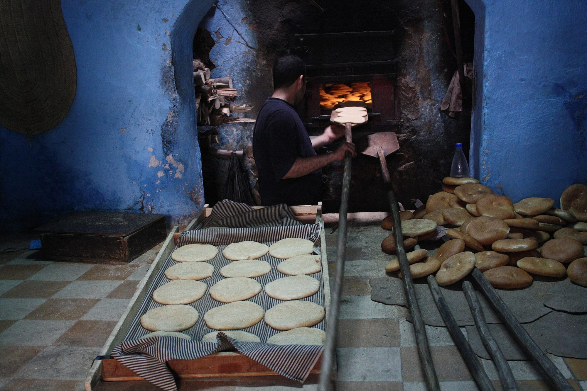 Photograph of a man baking round loaves of bread. The man is standing in front of an open oven built into a blue wall. He is holding a pan with a very long handle, preparing to slide it into the oven. The pan has three loaves of unbaked bread dough on it. A cart with many additional uncooked loaves is in the foreground. A pile of baked loaves is next to the oven.
