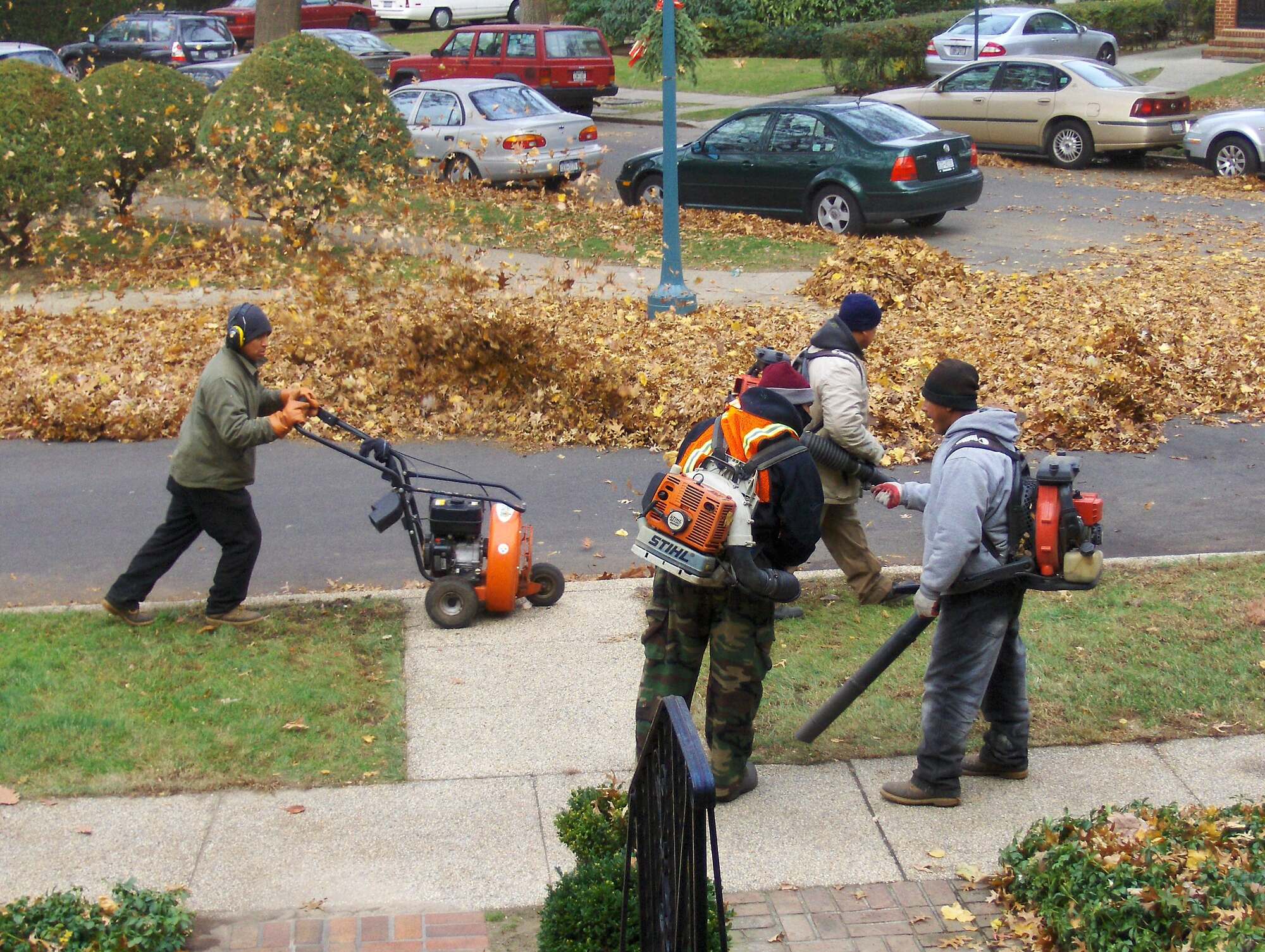 Photograph of four men doing yard maintenance. Three of the men are carrying leaf blowers, and one appears to be talking to another. A fourth man is pushing some other wheeled piece of lawn equipment.