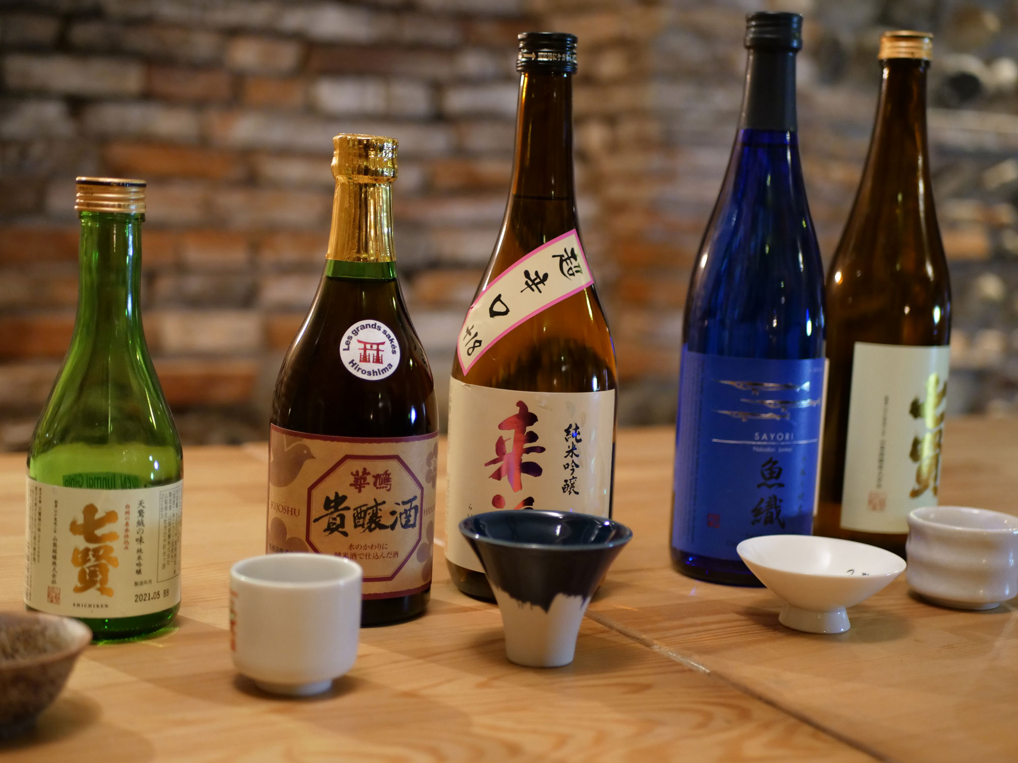 Photograph of bottles of sake with ceramic drinking cups in front of them. The sake bottles vary in height, color, shape, and label.