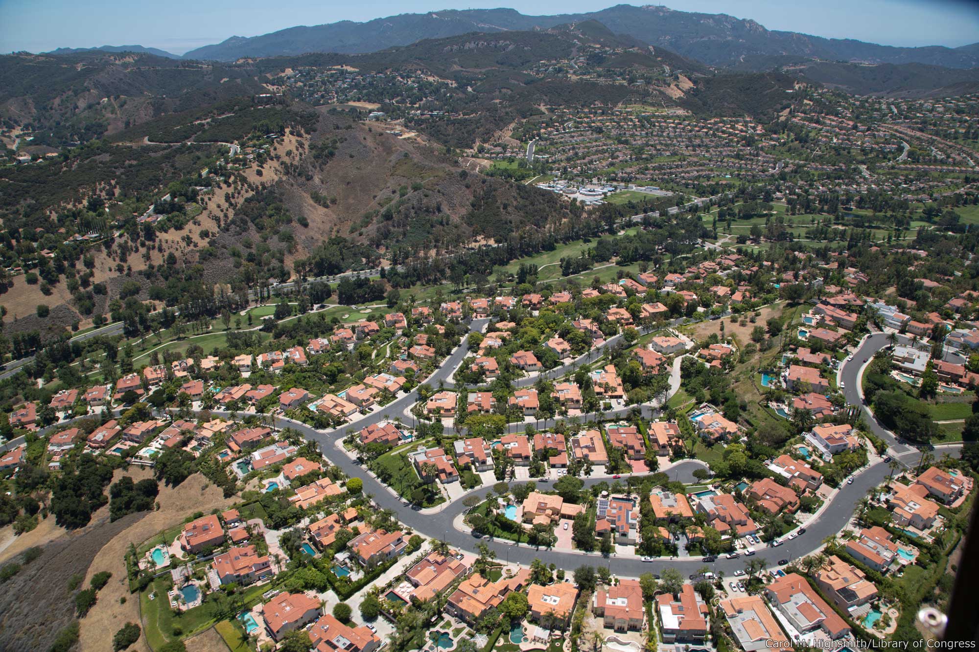 Aerial photograph of a housing development in Los Angeles, California, USA. The photo shows regularly spaced houses built along sinuous roads. The grass and trees around the houses are green. In the background, undeveloped hillsides can be seen. These have yellow and brown grass with scattered trees.