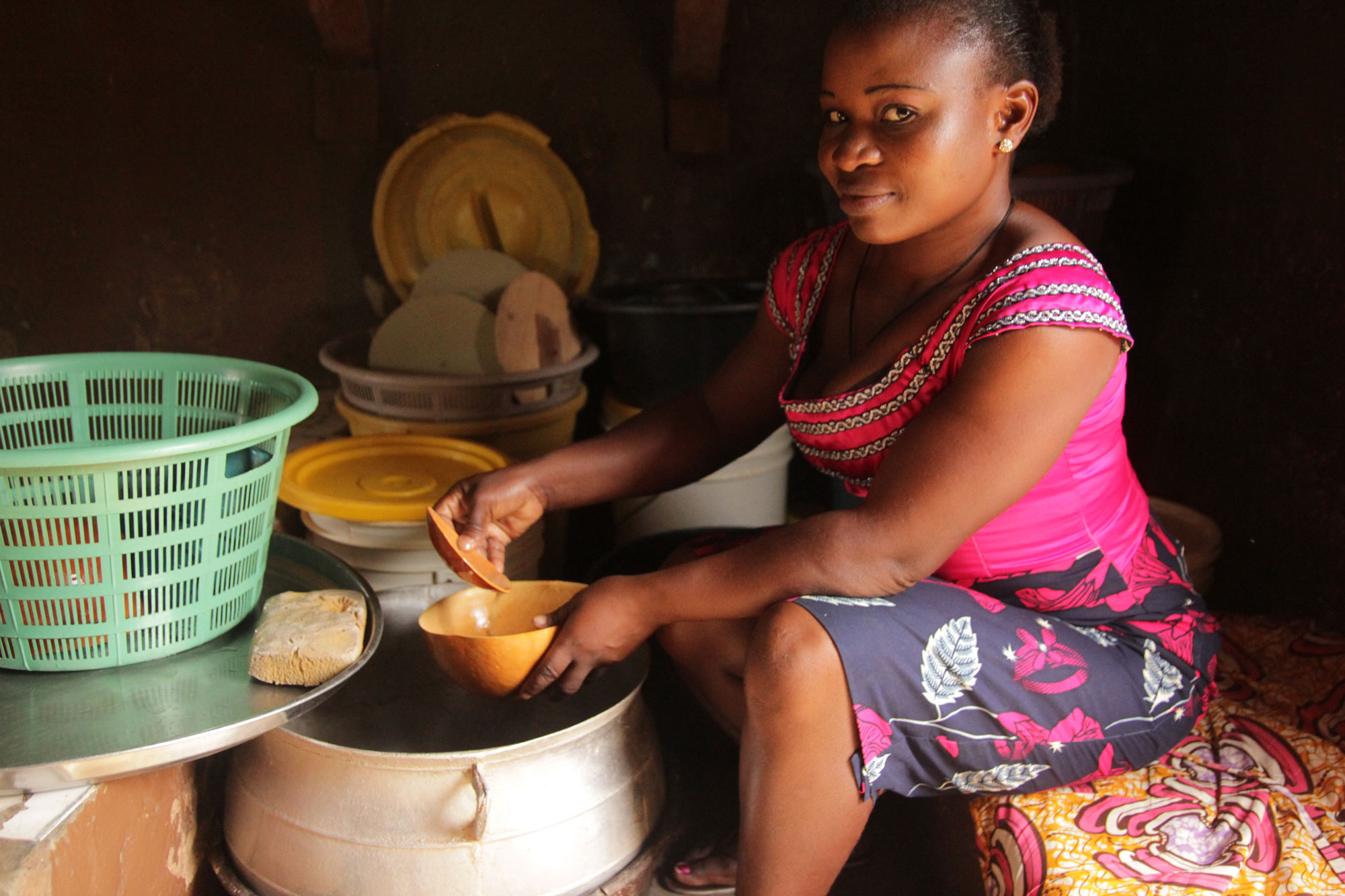 Photograph of a woman in Benin ladeling a traditional beer from a large kettle into a smaller bowl. The woman is seated and looking toward the camera.